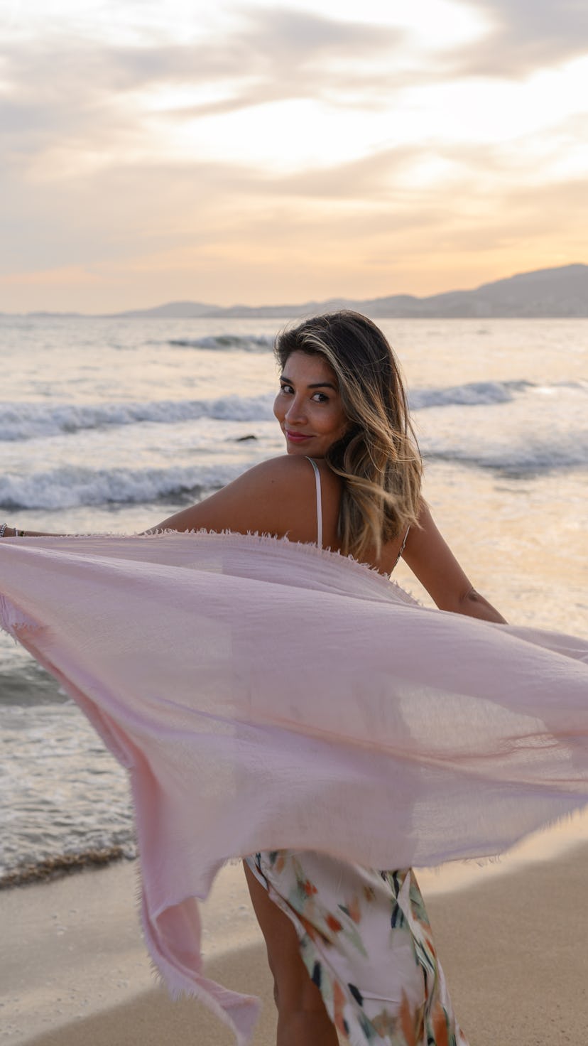 Beautiful young woman holding sarong. Female is enjoying sunset at weekend. She is at beach.