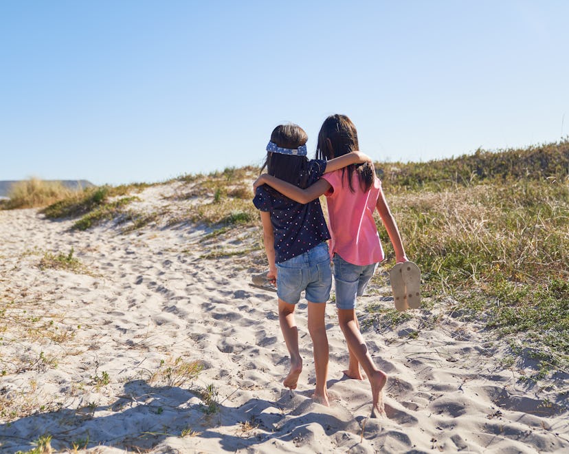 Happy, affectionate barefoot sisters walking on sunny, sandy beach, enjoying summer vacation