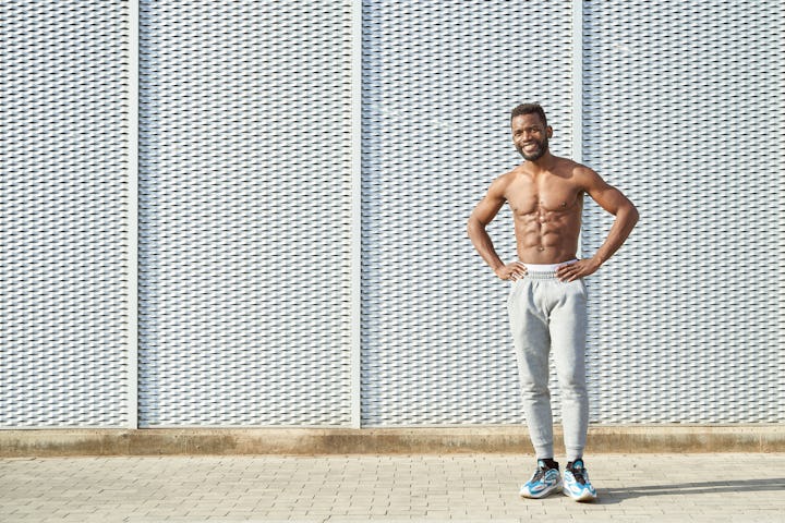 Shirtless African American muscular athlete standing against textured wall sweating after workout. S...