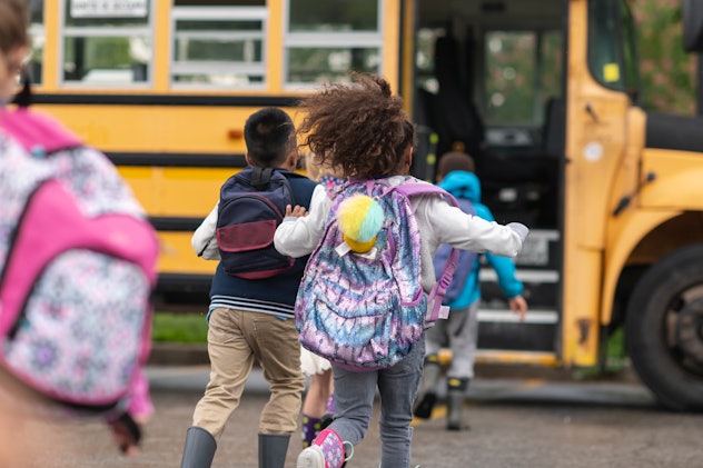 A group of elementary age children are getting on a school bus. The kids' backs are to the camera. T...