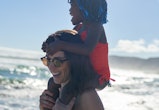 Smiling woman wearing sunglasses carrying toddler on shoulders on sunny ocean beach, last minute sum...