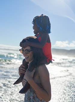Smiling woman wearing sunglasses carrying toddler on shoulders on sunny ocean beach, last minute sum...