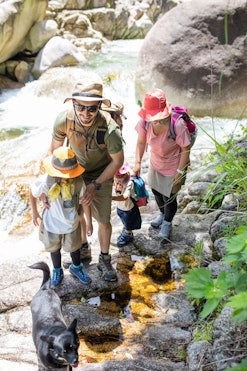 Two adult parents and two children wearing hats, carrying backpacks, hiking in a riverbed, demonstra...
