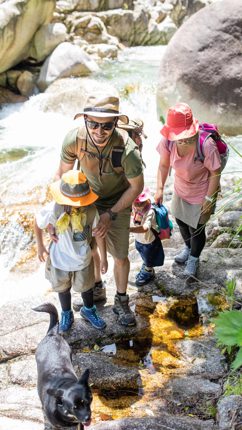Two adult parents and two children wearing hats, carrying backpacks, hiking in a riverbed, demonstra...