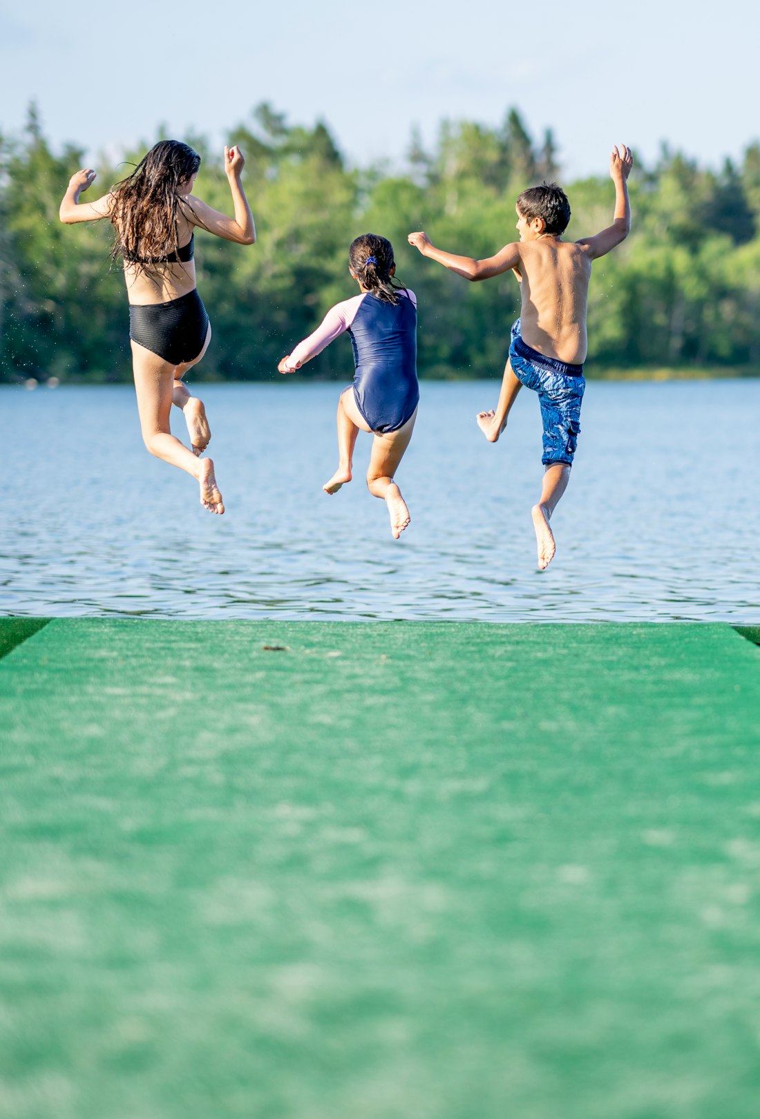 Three children in mid-air jumping off the end of a dock on a sunny summer day, last minute summer va...