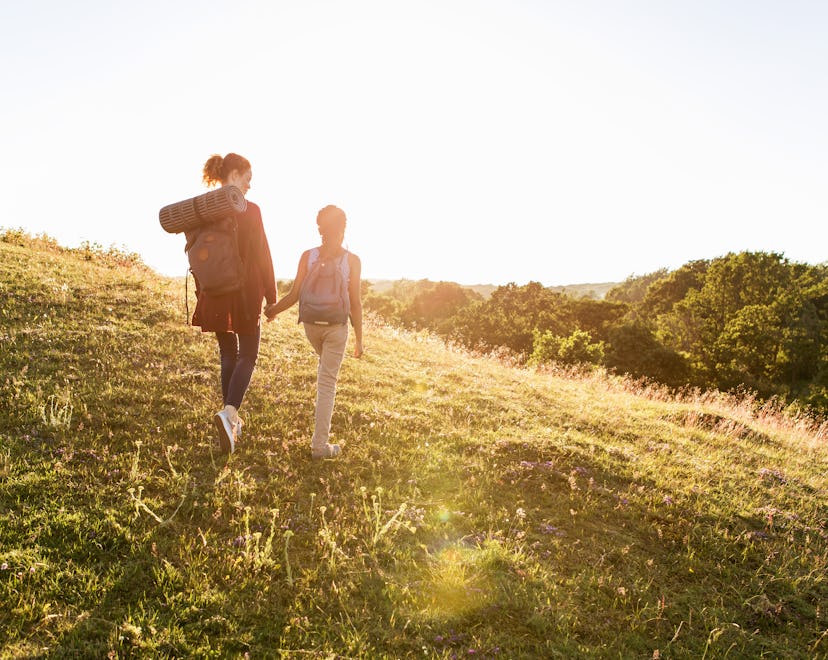 An adult and child wearing backpacks, holding hands, walking on a hill, facing away from camera with...