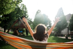 Back view of a girl in a sunny backyard sitting in hammock with hands in the air with a swing set in...