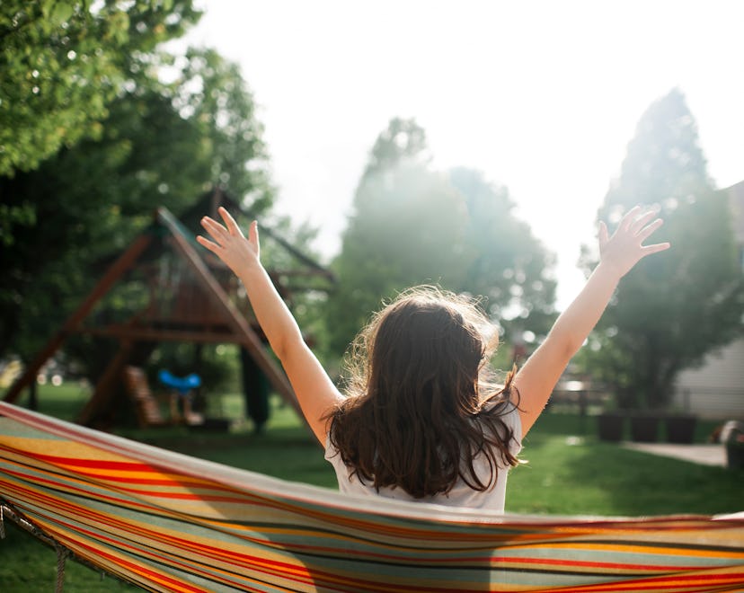 Back view of a girl in a sunny backyard sitting in hammock with hands in the air with a swing set in...