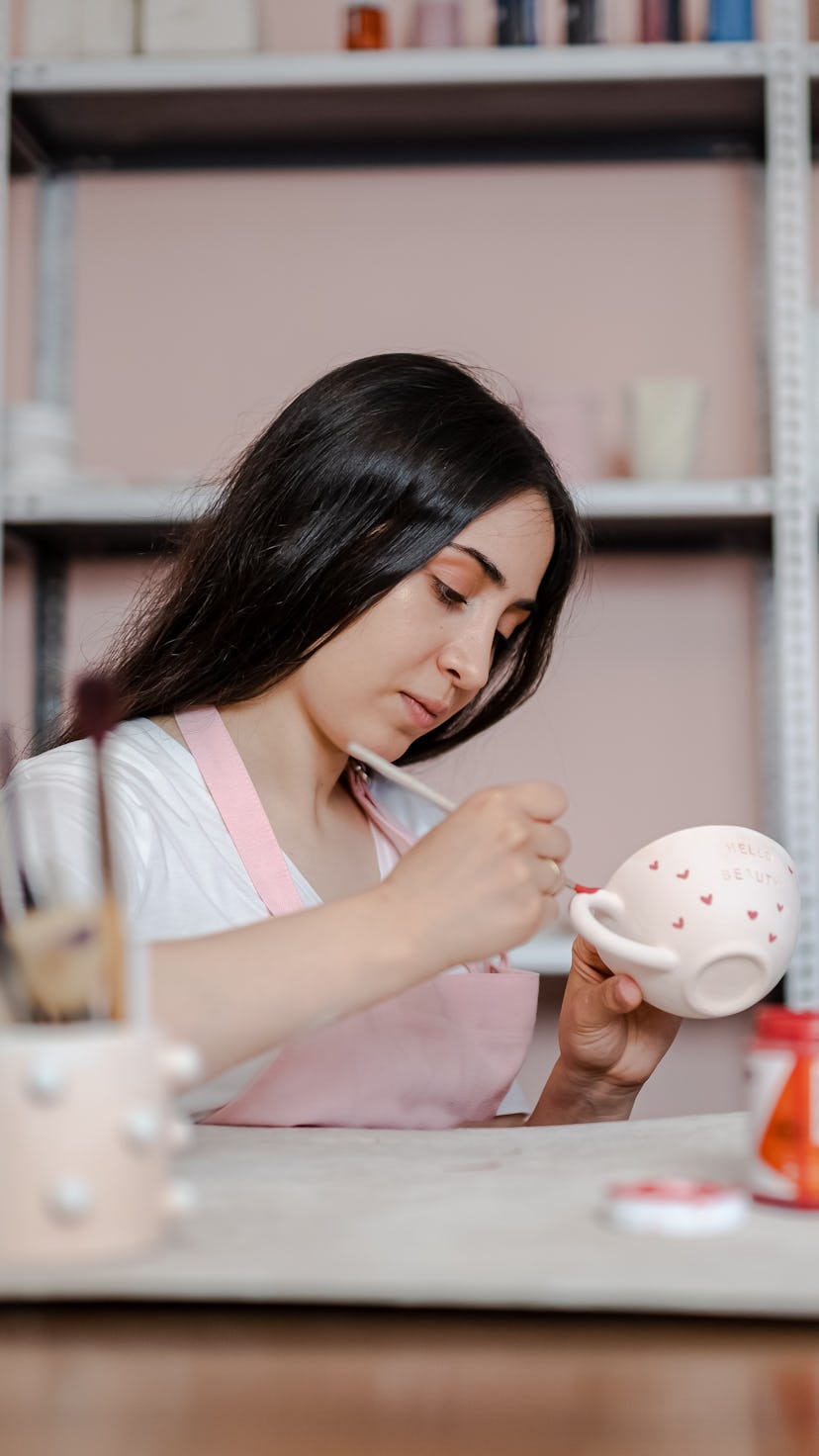 young woman is painting a ceramic mug in workshop. The July 2022 full moon is in the enterprising si...