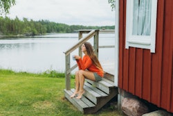 Young woman in red with long hair, sitting on the patio of the old wooden camping cabin drinking cof...