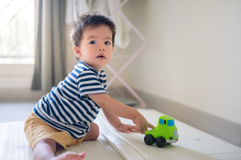 Adorable baby boy playing with his toy truck crawling on a soft play mat in the bedroom at home in a...