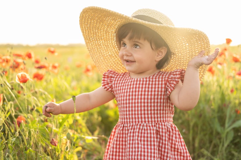 A toddler girl wearing a dress and floppy sun hat, smiling in a sunny field of flowers, in this arti...