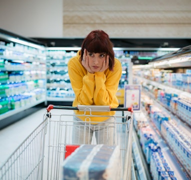 Young woman with red hair buying groceries in a local supermarket, feeling depressed by the high pri...