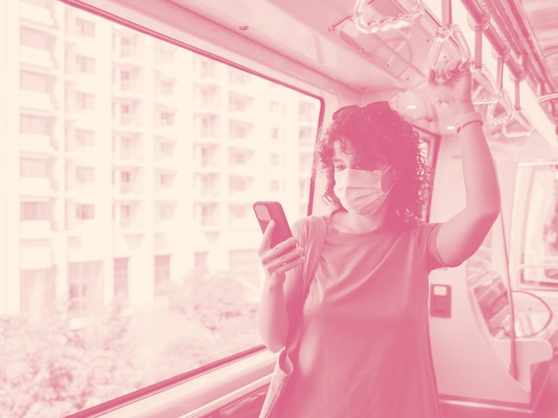 A young woman with curly hair riding the light rail wearing a face mask and checking her smartphone.