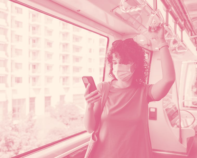 A young woman with curly hair riding the light rail wearing a face mask and checking her smartphone.
