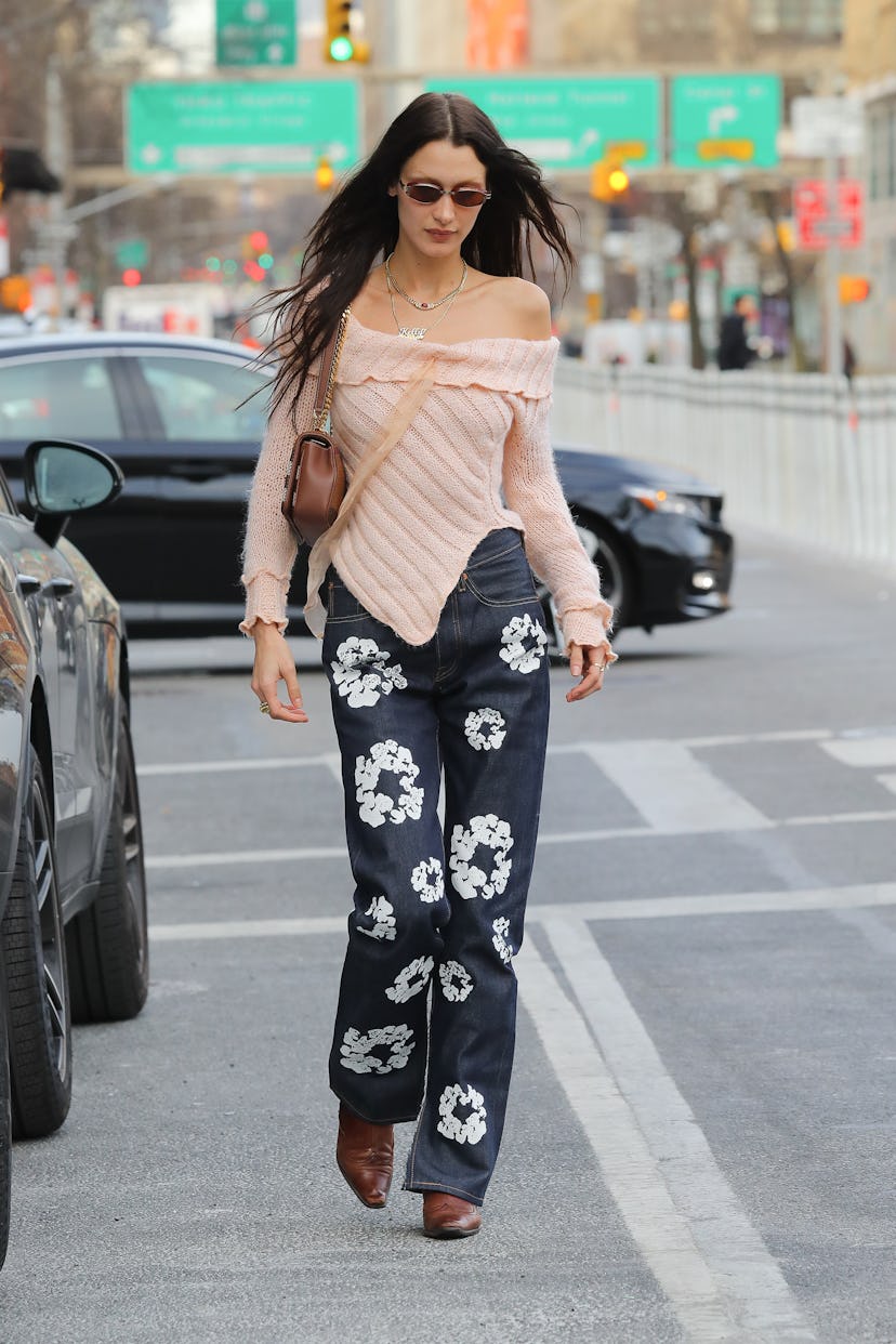 Bella Hadid wears brown cowboy boots with printed jeans and a light pink off-the-shoulder sweater on…