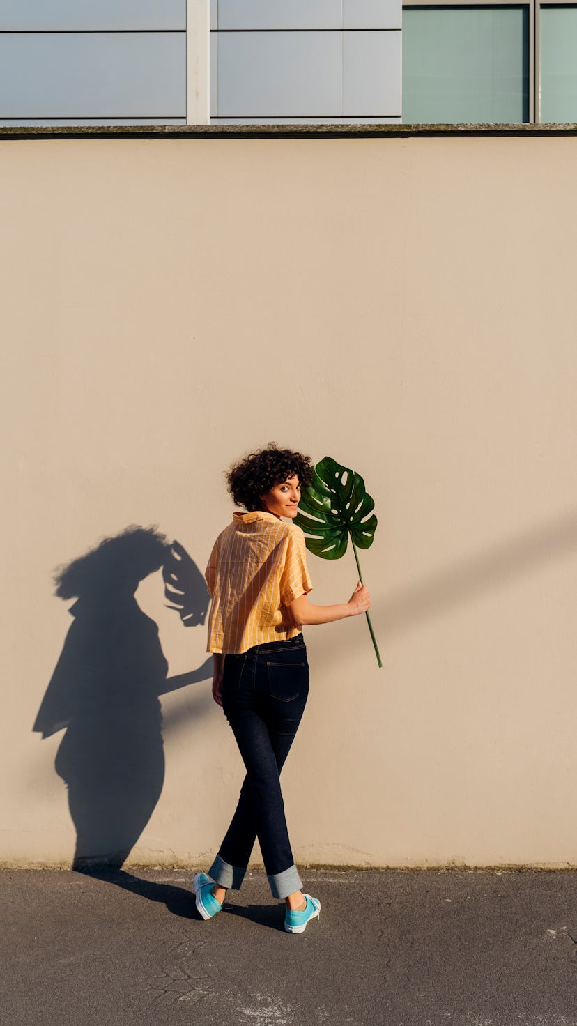 Woman holding a monstera plant against a wall, her shadow is behind her. Virgo season is about impro...