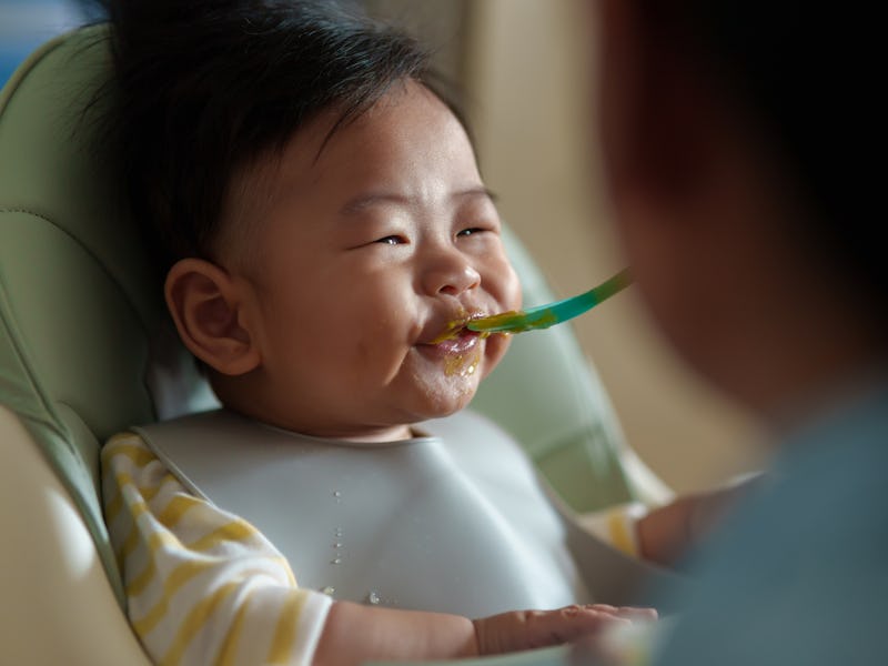 Cheerful Asian little baby boy in high chair enjoying while eating meal. Mother feeding cute baby wi...