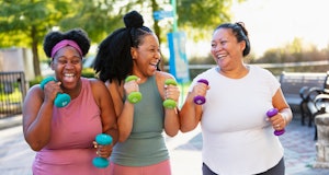 A multiracial group of three women exercising together in a city park. They are power walking with h...