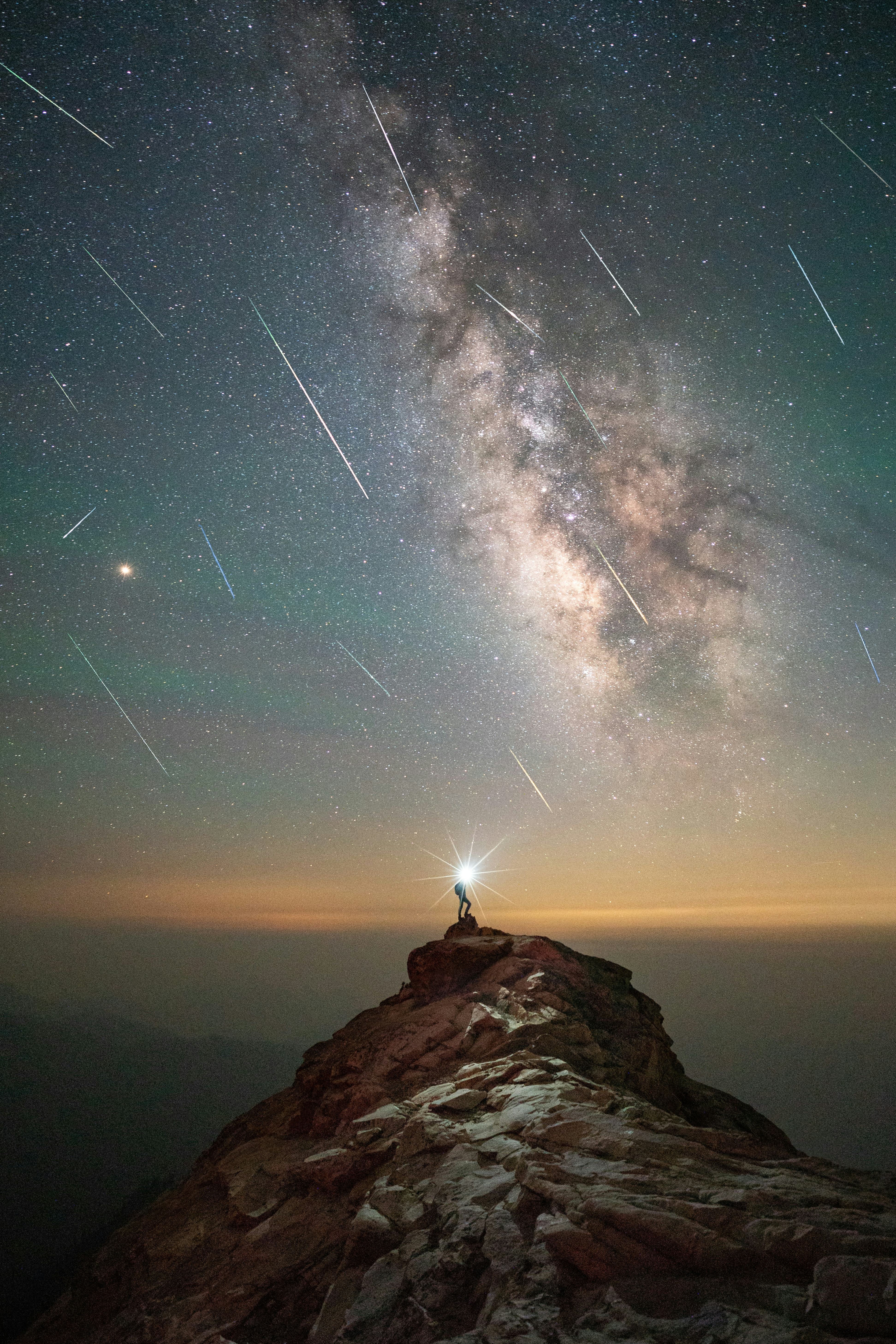 A person stands on a cliff with the Milky Way visible in the background