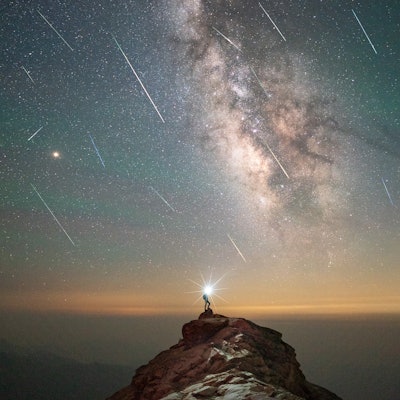 A person stands on a cliff with the Milky Way visible in the background