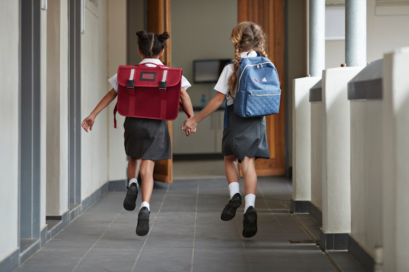 Two girls with their backpacks on, holding hands while skipping through the hallway in school.