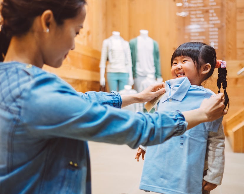 Pretty young mom fitting a shirt on the cheerful little girl at the kid's wear department in a fashi...