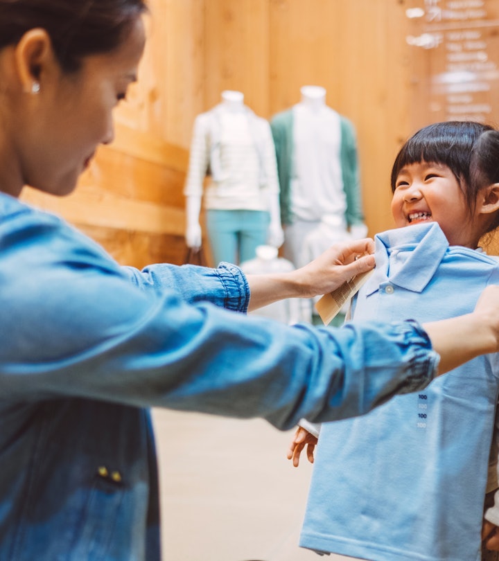 Pretty young mom fitting a shirt on the cheerful little girl at the kid's wear department in a fashi...