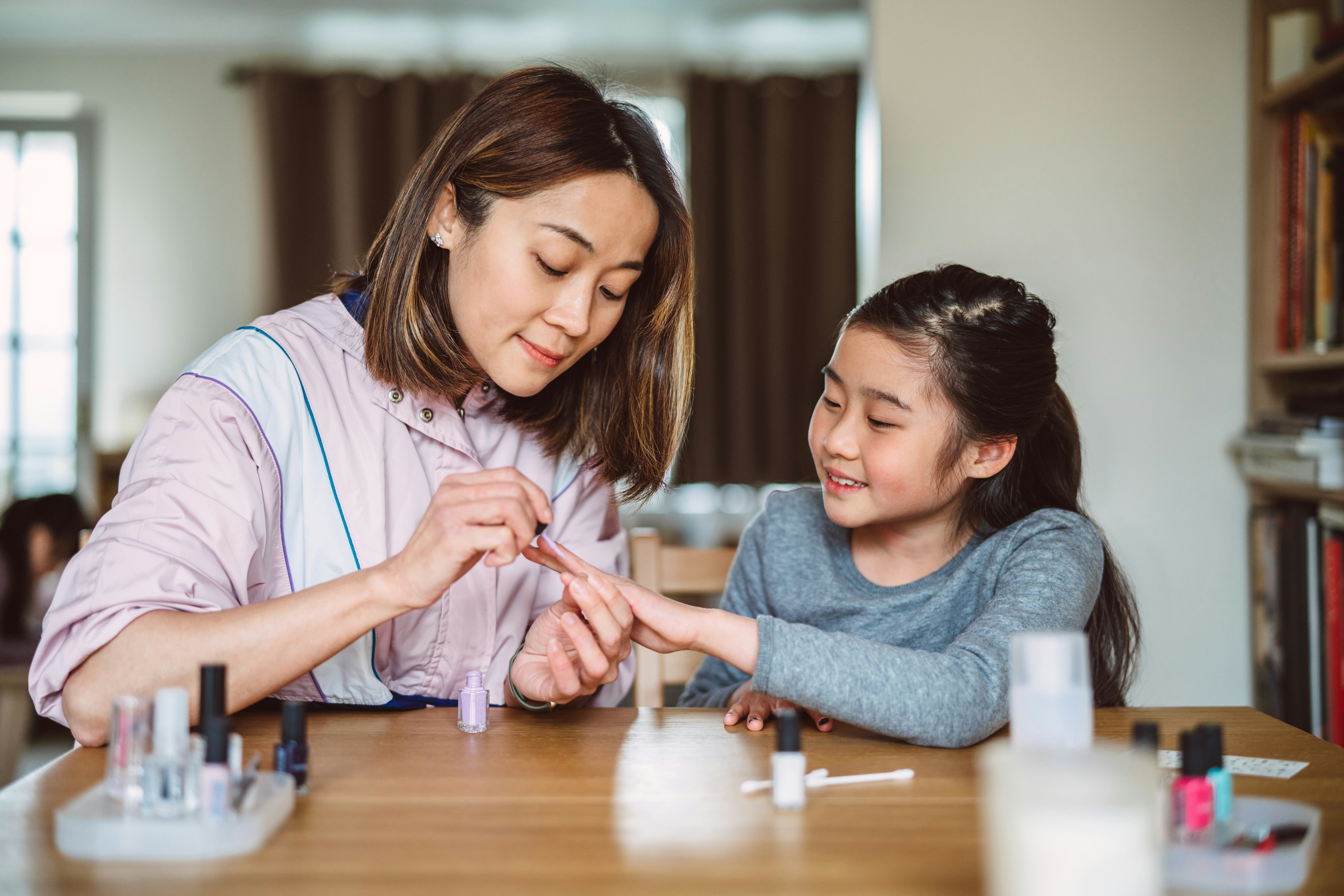 Young pretty mom polishing her lovely daughter’s fingernails at home joyfully.