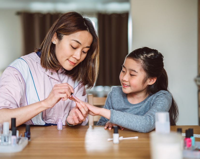 Young pretty mom polishing her lovely daughter’s fingernails at home joyfully.