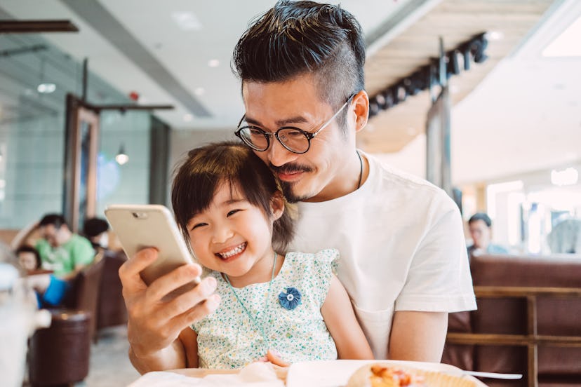 Young dad using smartphone with his little daughter while little daughter sitting on his lap in a co...