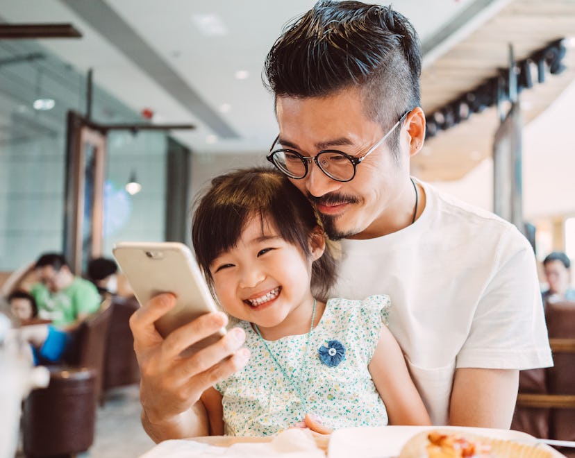 Young dad using smartphone with his little daughter while little daughter sitting on his lap in a co...