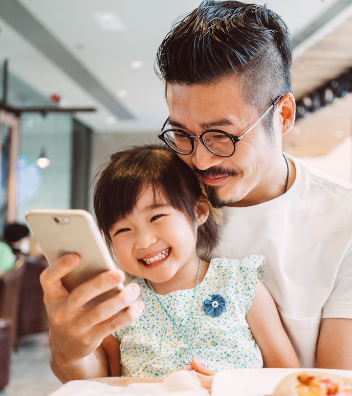 Young dad using smartphone with his little daughter while little daughter sitting on his lap in a co...