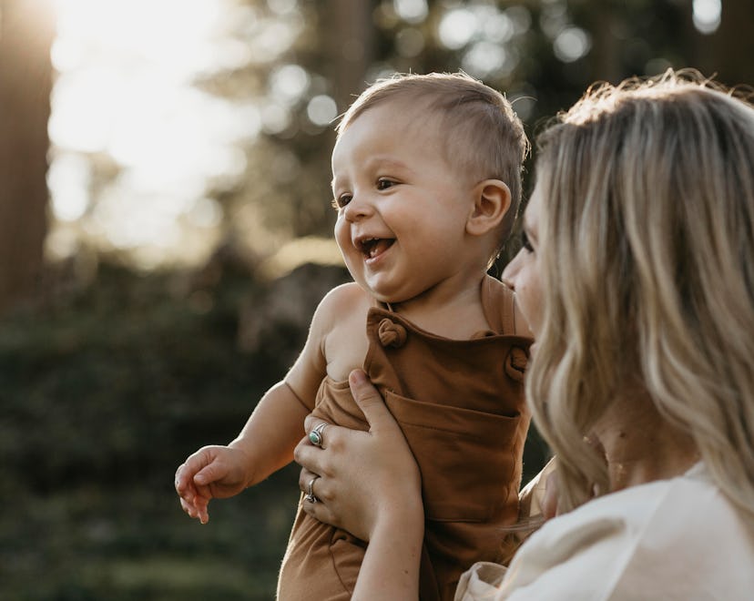 A mother holding a smiling toddler boy wearing a brown romper