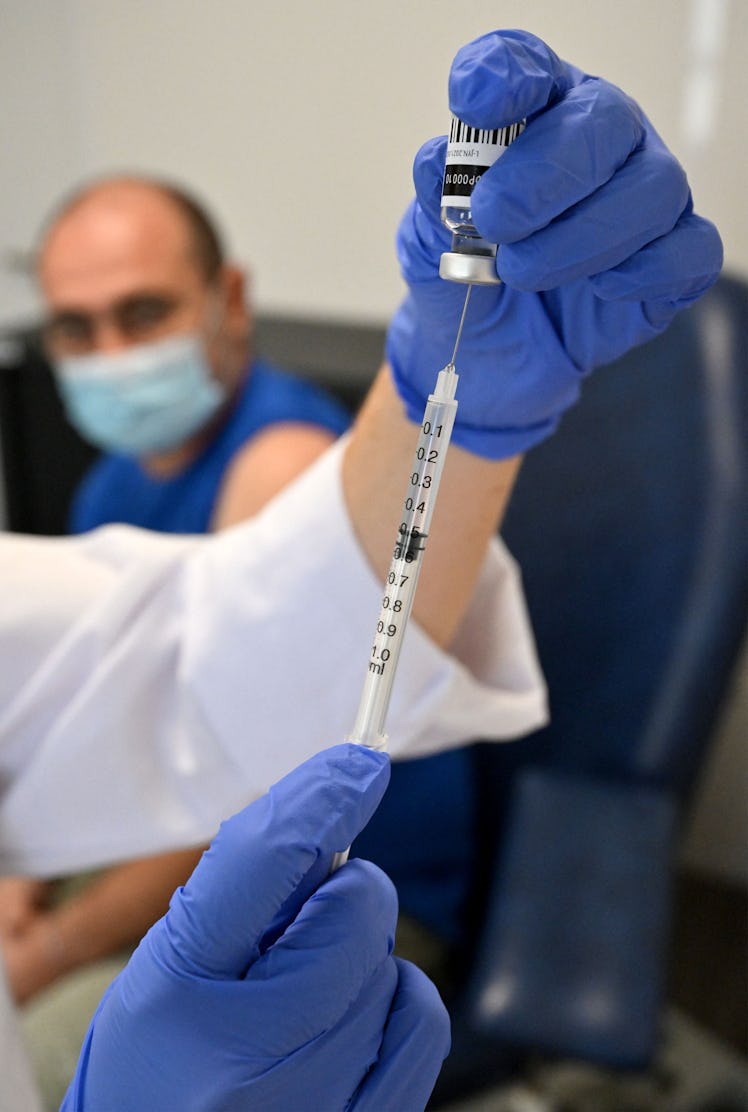 A nurse prepares a vaccine with a man in a mask waiting in the background to receive it