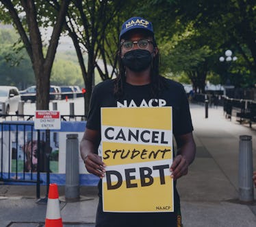 A protestor with a mask on and a hat holds a sign that says "Cancel Student Debt"