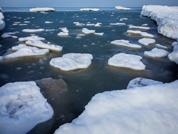 Melting ice blocks floating in dark sea water with frozen shore under cloudy blue sky in sunny weath...