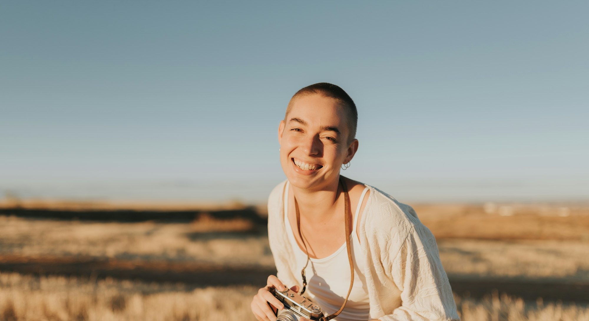 Woman with short hair looking at camera and laughing loudly holds vintage camera at sunset. She is w...