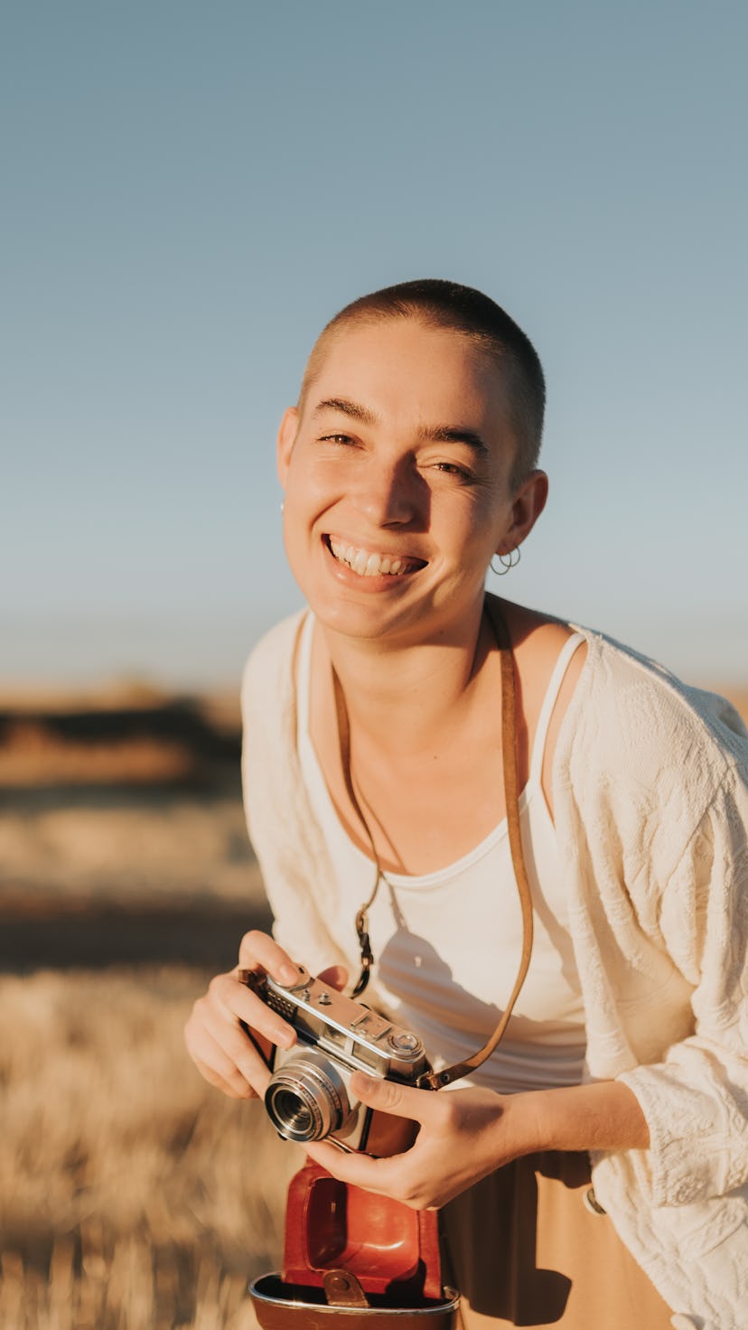 Woman with short hair looking at camera and laughing loudly holds vintage camera at sunset. She is w...