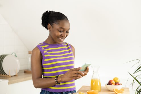 Portrait of a happy young woman making fresh juice and using smartphone at her kitchen