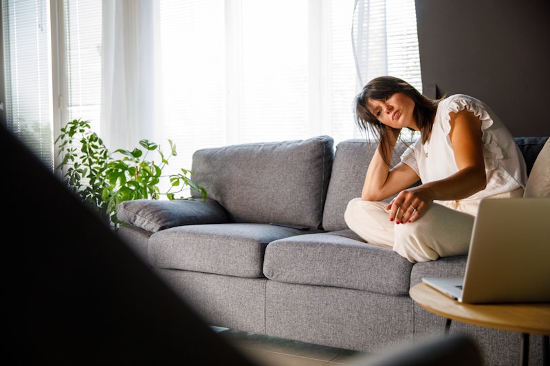 Wide shot of distraught mid adult woman sitting on the sofa in her living room, head in hand, lookin...
