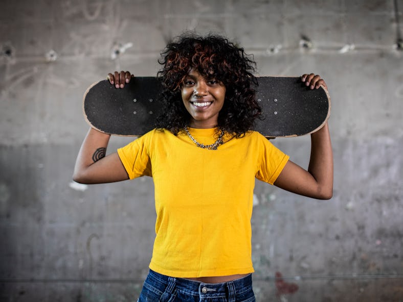 Girl with shag haircut posing in a yellow shirt while holding a skateboard 
