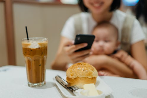 An Asian Chinese woman carrying her baby boy and taking photo of her dessert on table with smartphon...