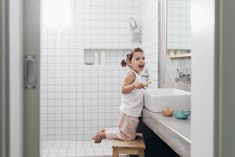 Toddler kneeling on a stool at a bathroom sink, holding a toothbrush, with mouth open to the camera,...