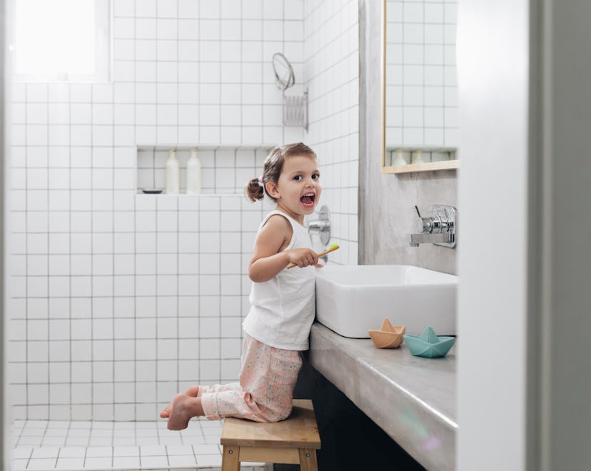Toddler kneeling on a stool at a bathroom sink, holding a toothbrush, with mouth open to the camera,...