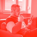 Man relaxing on the sofa with electronic cigarette in mouth, holding smart phone and scrolling throu...