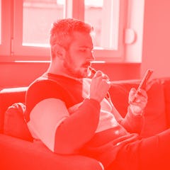 Man relaxing on the sofa with electronic cigarette in mouth, holding smart phone and scrolling throu...