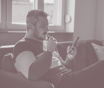 Man relaxing on the sofa with electronic cigarette in mouth, holding smart phone and scrolling throu...