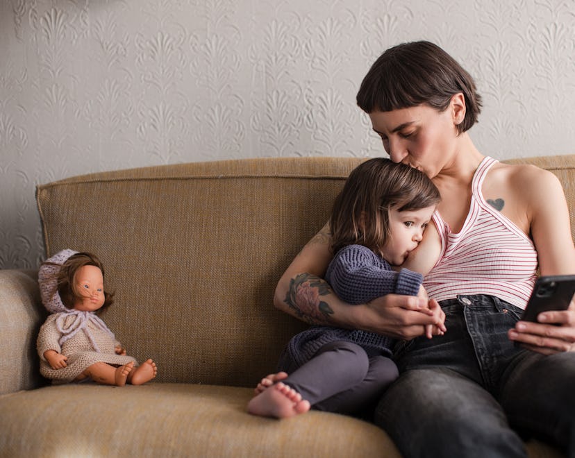 Beautiful young mom kissing and extended breastfeeding her toddler on the couch and holding her phon...