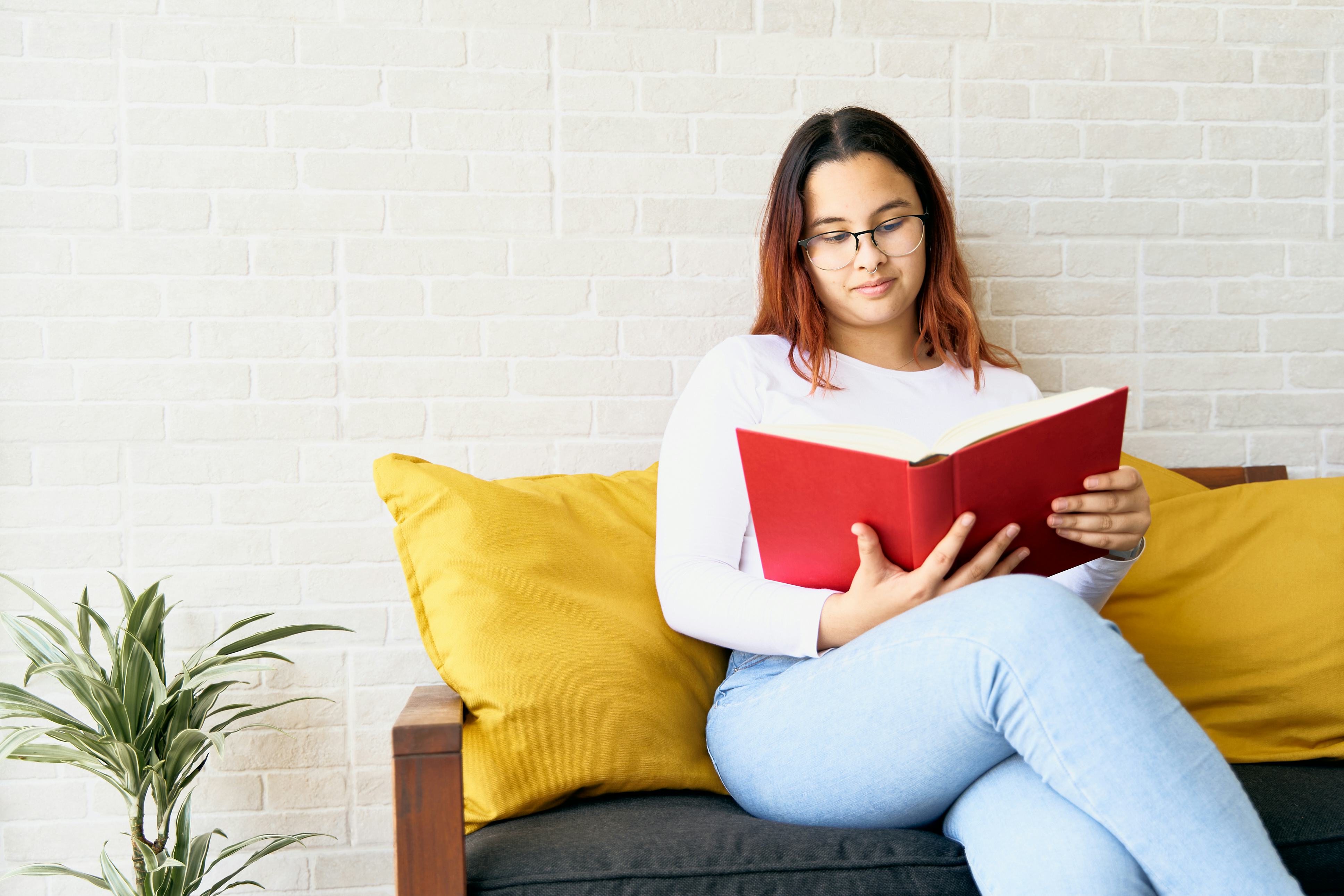 Beautiful teenage girl enjoying reading red book at home sitting on sofa with yellow cushions. She h...